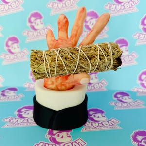 May include: A unique sculpture featuring a bundle of dried sage held by a hand-shaped form. The hand is painted with red accents, and the sage is secured with white string. The sculpture rests on a white and black cylindrical base. The background is a light blue pattern with the text "LUB ART".