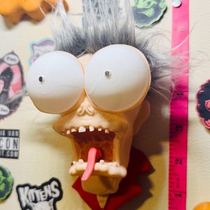 May include: A novelty fridge magnet shaped like a cartoon character with large white eyes, grey hair, and an open mouth with a pink tongue. The magnet is attached to a surface with other magnets and stickers. A pink measuring tape is visible.