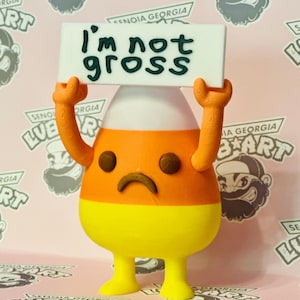 May include: A 3D-printed candy corn figure with orange arms and a sad face holds a sign that reads "I'm not gross." The figure is yellow, orange, and white. The background is pink with repeating logos.