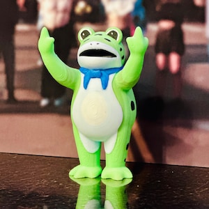 May include: A green frog figurine with a blue bow tie, black spots, and white belly. The frog is raising its arms with the middle finger extended. The figurine is standing on a reflective surface.