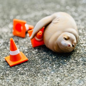 May include: A small, beige, rounded figurine resembling a seal with a smooth texture, positioned next to three miniature orange traffic cones with white reflective bands. The figurine has a soft, rounded body, a small snout, and closed eyes.