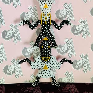 May include: A whimsical sculpture featuring two figures stacked on top of each other. The figures are white with black polka dots and have outstretched arms. Yellow accents include a triangle and facial features. The background is pink with repeating text.