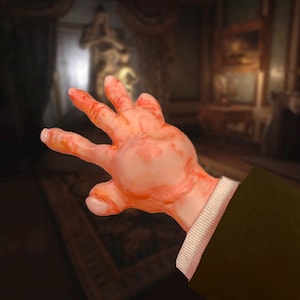 Può includere: Primo piano di una mano coperta di liquido rosso, con una manica verde scuro e un polsino bianco. La mano è in un gesto, con le dita divaricate. Lo sfondo è un interno sfocato.