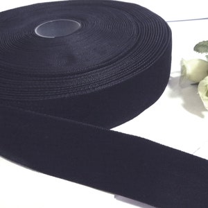 May include: A roll of black velvet ribbon. The ribbon is wide and has a soft, plush texture. The roll is tightly wound, and a length of ribbon extends from the roll. The background is white with a few white flowers.