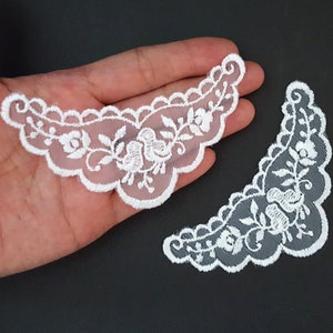 Könnte beinhalten: Zwei weiße Spitzenapplikationen mit Blumenmuster. Die Applikationen sind halbmondförmig und haben einen gewellten Rand.