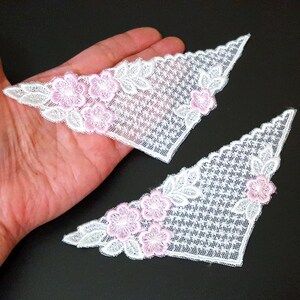 May include: Two white lace fabric corner patches with pink flower embroidery. The patches are triangular in shape and have a delicate, intricate design.