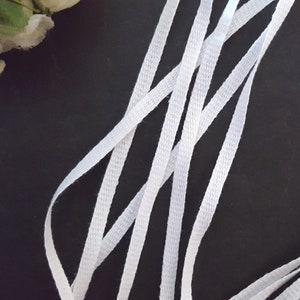 May include: White cotton shoelaces, flat and woven, arranged in a criss-cross pattern on a black background.