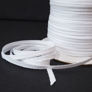 May include: A spool of white cotton twill tape. The tape is approximately 1/4 inch wide and is wound around a cardboard spool.