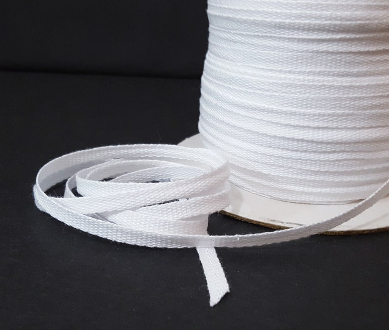 May include: A spool of white cotton twill tape. The tape is approximately 1/4 inch wide and is wound around a cardboard spool.