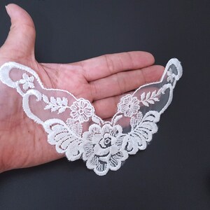May include: A white floral lace applique with an openwork design. The applique features a central rose surrounded by smaller flowers and leaves. The lace is held in a hand against a dark background, showcasing its intricate details.