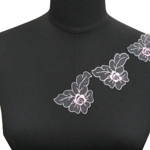 Könnte beinhalten: Drei weiße Blumenapplikationen mit rosa Zentren auf schwarzem Hintergrund. Die Applikationen sind aus Spitze und eignen sich perfekt, um jedem Projekt einen Hauch von Eleganz zu verleihen.