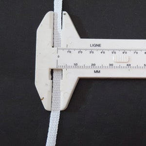 May include: A white measuring tool with a white fabric ribbon in the jaws. The tool has markings in inches and millimeters. The markings read "LIGNE" and "MM".