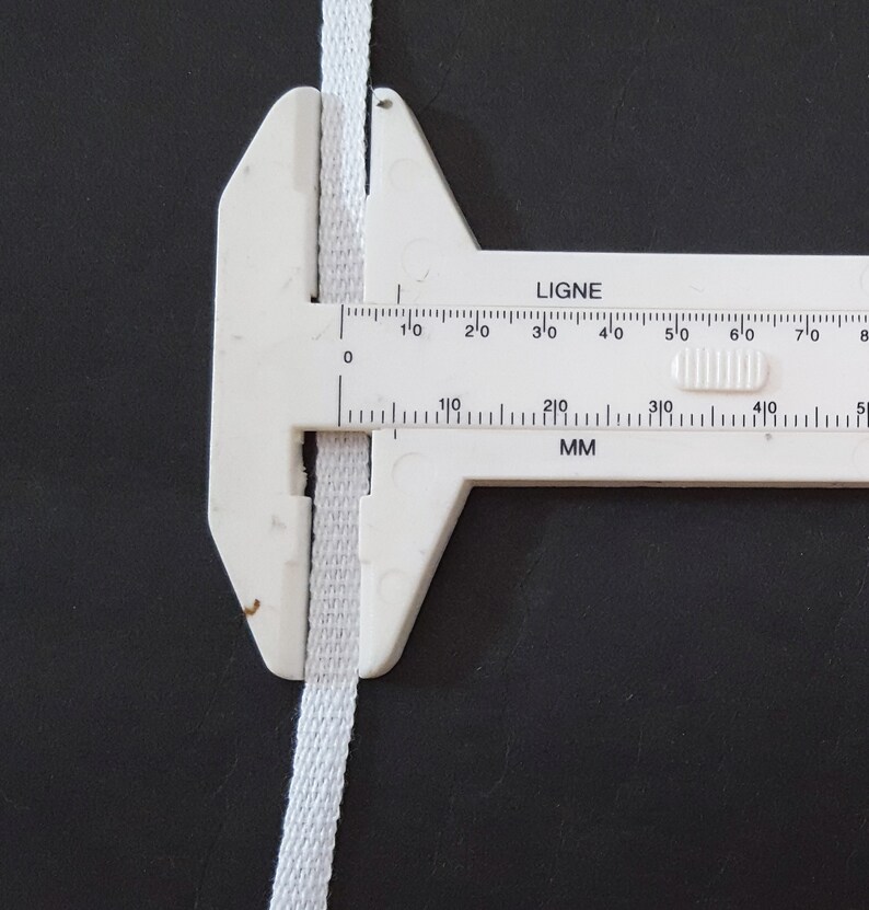 May include: A white measuring tool with a white fabric ribbon in the jaws. The tool has markings in inches and millimeters. The markings read "LIGNE" and "MM".