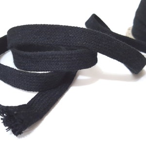 May include: Black cotton cord, a thick, woven fabric ribbon, is coiled on a white background. The cord is approximately 1.2 cm wide.