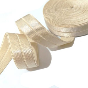 May include: A roll of beige, ribbed elastic ribbon. The ribbon is folded and has a central seam. The roll is partially unraveled, with a length of the ribbon extending out. The background is white.