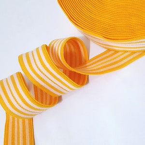 Może przedstawiać: Elastyczna lamówka w paski żółte i białe. Lamówka jest złożona i zwinięta, co ukazuje wzór w paski.