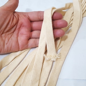 May include: Close-up of several cream-coloured, flat cotton ribbons. The ribbons are woven with a textured pattern and have a frayed edge. The ribbons are held in a hand, against a white background.