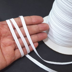 May include: White cotton cord, approximately 1/4 inch wide, is shown in a hand and on a spool. The cord is a flat, woven type of cord.