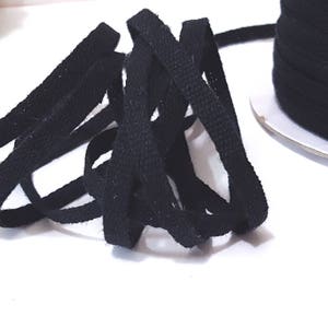 May include: Black cotton cord, a type of flat, woven ribbon, is shown in a loose pile. The cord is approximately 1/4 inch wide and is available in a spool.