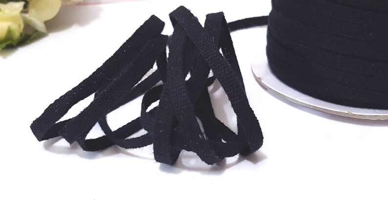 May include: Black cotton cord, a type of flat, woven ribbon, is shown in a loose pile. The cord is approximately 1/4 inch wide and is available in a spool.