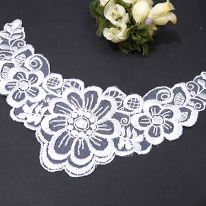 May include: White lace trim with a floral design. The trim features a large flower in the centre with smaller flowers and leaves surrounding it.