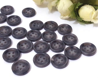 May include: A collection of 18 dark gray plastic buttons with four holes each. The buttons are arranged in a random pattern on a white surface.