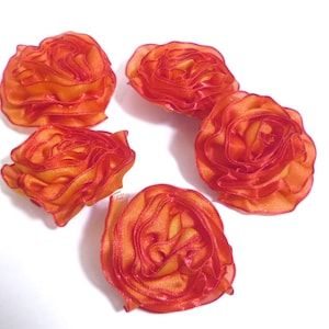 Könnte beinhalten: Fünf orangefarbene Stoffrosen-Verzierungen mit Rüschenkanten sind auf einer weißen Oberfläche angeordnet. Die Rosen haben ein geschichtetes Design mit einem Farbverlauf aus Orange- und Gelbtönen. Diese dekorativen Blumen eignen sich für verschiedene Bastelprojekte.