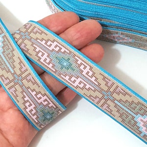 May include: A turquoise blue ribbon with a beige and pink geometric pattern. The ribbon is woven with a repeating design of squares and diamonds.