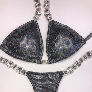 Puede incluir: Conjunto de bikini de dos piezas en tejido gris oscuro brillante. La parte superior e inferior están adornadas con pedrería rectangular transparente a lo largo de las correas y la cintura. El bikini tiene un top triangular clásico y una braguita tipo tanga.