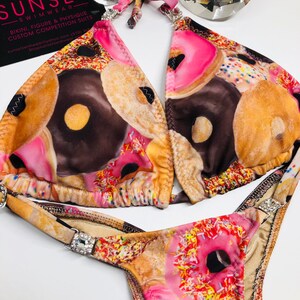 Puede incluir: Un bikini rosa, marrón y blanco con estampado de donuts. La parte superior del bikini es de estilo halter con cuello en V y la parte inferior es de estilo de corte alto. El bikini está decorado con diamantes de imitación.