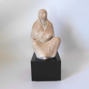 Op de afbeelding: Een keramisch beeld van een zittende vrouw die een sjaal draagt. Het beeld is geschilderd in tinten bruin en wit en wordt getoond op een zwarte basis.