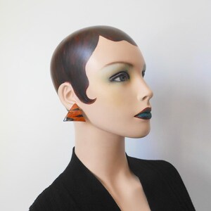 Op de afbeelding: Een mannequin draagt een zwarte top en een paar geometrische driehoekige oorbellen. De oorbellen hebben een patroon van oranje en zwarte strepen. De oorbellen zijn een opvallend sieraad.