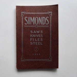Può includere: Un libro rilegato in pelle rossa con il titolo "Simonds" e il testo "Saws Knives Files Steel" in lettere argentate. Il libro ha anche l'anno "1923" stampato sulla copertina.