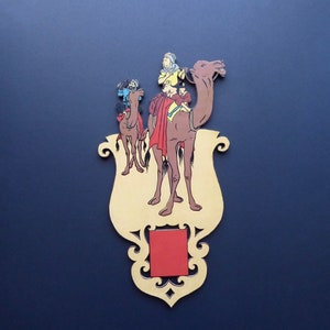 May include: A decorative wooden plaque featuring a camel and three riders. The camel is brown with a yellow saddle and the riders are wearing colourful clothing. The plaque is painted in a vintage style and has a red square in the centre.