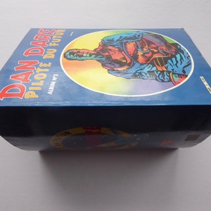 May include: A blue and yellow comic book titled "Dan Dare Pilote du Futur" with the text "Album N&deg;2" on the cover. The cover features a colourful illustration of a man in a blue jumpsuit and helmet.