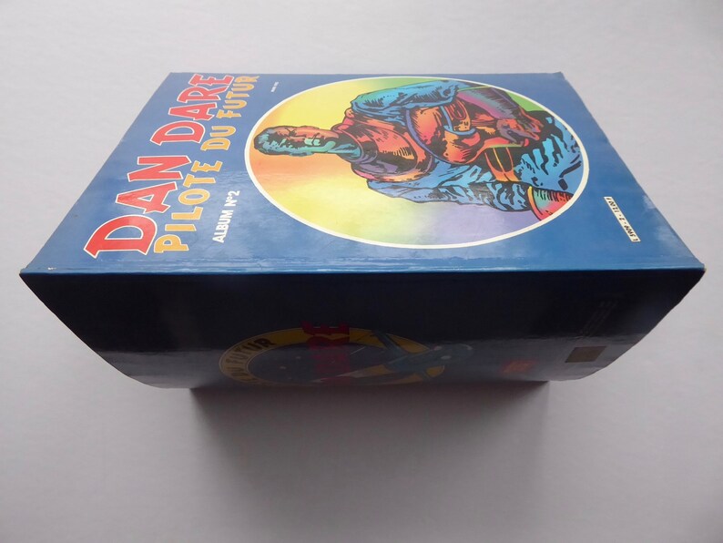 May include: A blue and yellow comic book titled "Dan Dare Pilote du Futur" with the text "Album N&deg;2" on the cover. The cover features a colourful illustration of a man in a blue jumpsuit and helmet.