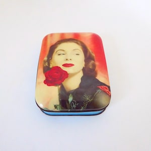 Può includere: Una scatola di latta in stile vintage con un'immagine colorata di una donna con rossetto rosso e una rosa rossa. Lo sfondo è un gradiente rosso e arancione.