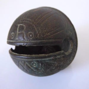 May include: A dark bronze-colored, spherical bell with an open slit. The bell has etched lines and the letter "R" on its surface. The bell's design suggests it may be an antique or vintage item.