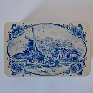 May include: A white and blue tin with a Dutch windmill scene and the word "Verkade" printed on the bottom. The tin features a blue and white windmill, trees, and a house.
