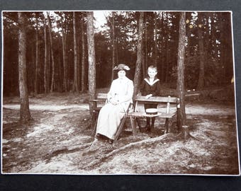 Photo of woman and child. Black and white glossy paper 1910. Woman sitting on bench. Forest. Sailor suit. Sailor hat. Flower hat. Cane