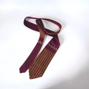 May include: A burgundy tie with a geometric pattern in gold and brown. The tie is made of a smooth fabric and features a repeating triangle design on the lower portion and zigzags near the knot.