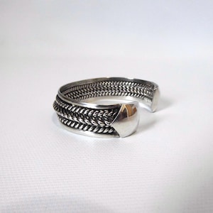 Op de afbeelding: Een zilveren manchetarmband met een gevlochten ontwerp. De armband heeft een dikke band van geweven metaal, met een afgerond, gepolijst uiteinde. Het open ontwerp maakt het gemakkelijk om te dragen. De sieraden worden op een wit oppervlak gepresenteerd.