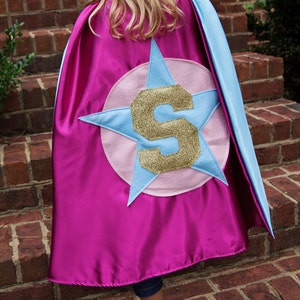 May include: A magenta superhero cape with light blue trim and a light blue starburst design. The starburst has a pink circle in the center with a gold glitter letter "S".