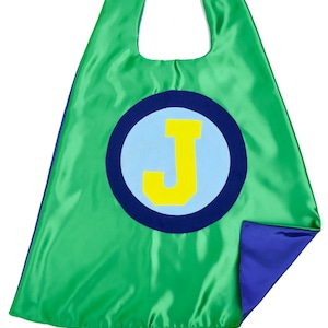 May include: A reversible superhero cape in green and blue. The green side features a large blue circle with a light blue center and a yellow letter "J". The cape has a hook and loop closure at the neck.