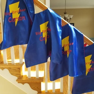 May include: Blue superhero capes with yellow lightning bolts and the words "Big Brother" followed by a name in red. The capes are draped over a wooden staircase railing.