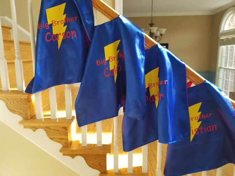 May include: Blue superhero capes with yellow lightning bolts and the words "Big Brother" followed by a name in red. The capes are draped over a wooden staircase railing.