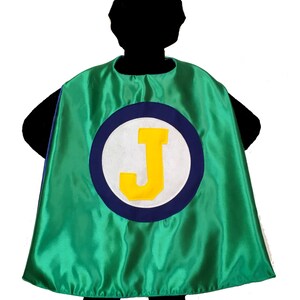 May include: A green satin superhero cape with a large circular emblem. The emblem features a white circle with a blue border and a yellow letter "J". The cape is designed to be worn by a child.