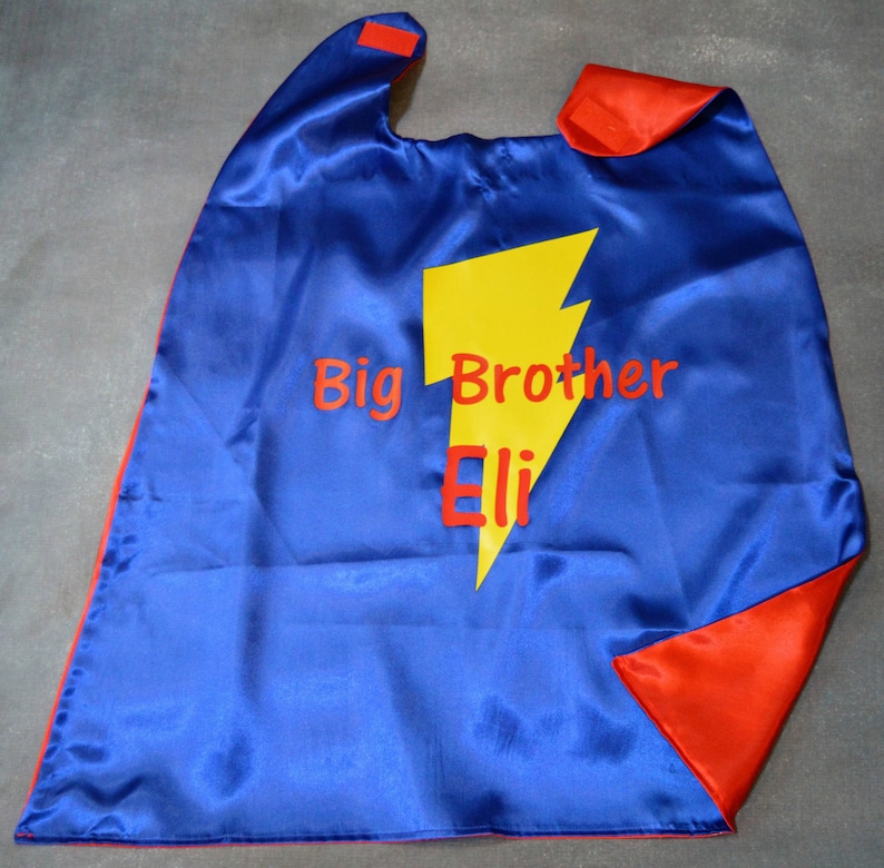 May include: A reversible blue and red satin superhero cape with the words "Big Brother Eli" and a yellow lightning bolt graphic. The cape has a hook and loop closure at the neck.