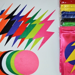 May include: A collection of colorful felt cutouts and flags. The felt cutouts include lightning bolt shapes in various colors like blue, orange, black, purple, red, green, and pink. There are also circles in multiple colors. The flags are pink with a blue circle and yellow lightning bolt.