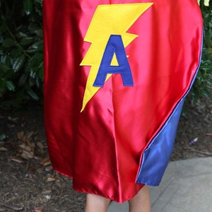 May include: A red superhero cape with a yellow lightning bolt and a blue letter "A" applique. The cape has a blue lining and is worn by a child. The cape is made of a shiny, satin-like material.